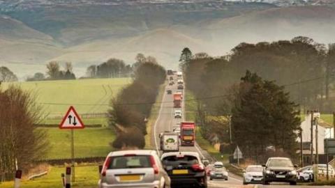 A stretch of single-carriageway traffic on the A66. The road is busy with cars and lorries, there are green fields either side of the road and the fells are visible in the background.