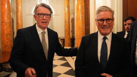 Lord Mandelson stands next to Keir Starmer. Both are wearing suits.