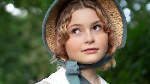 Theia looks off into the distance, wearing a blue trimmed bonnet and a white dress. She has blonde hair and brown eyes and is wearing subtle make-up.