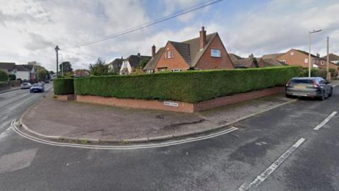 Entrance to Briar Close, Exmouth. There is a main road to the left with a smaller road with a large hedge at the entrance. A small brick wall has a sign that reads "Briar Close" on it. 