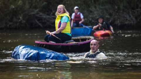One man with balding hair is pictured in the water next to a capsized coracle. A woman in a yellow life jacket remains on her craft behind. A third coracle and rider is in the background, alongside a man in a kayak.