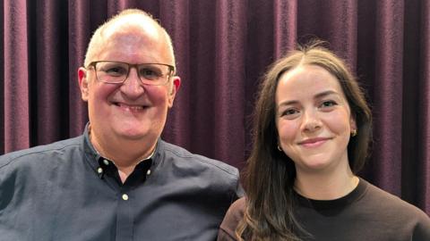 Spencer and Hollie stand side by side in front of a purple curtain, smiling to the camera. Spencer is a middle-aged, balding man with glasses, wearing a dark blue shirt. Hollie is a young woman with long brown hair, wearing a brown top.