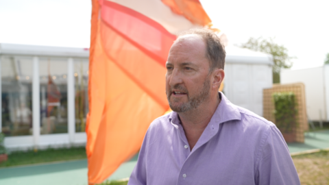 Guto Harri is facing sideways to the camera, wearing a lavender shirt, stood on the Hay Festival grounds. 