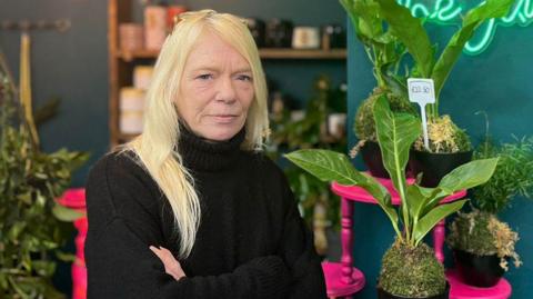 A blonde woman in a black polo-neck jumper stands inside her shop surrounded by plants, some placed on pink tables.  