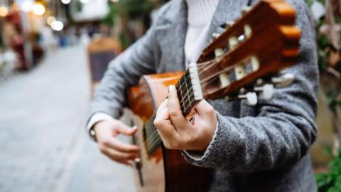 Generic image of someone playing a guitar at the side of a pedestrianised street with a blurred background of premises with lights showing. They are wearing a grey jacket and white polo-neck. 
