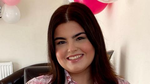 Ayla Jones is a white young woman with shoulder length dark brown hair. She is wearing a floral pink blouse. Behind her on the wall are some clear, white and pink balloons and there is pink confetti on the table. She is smiling directly at the camera. 