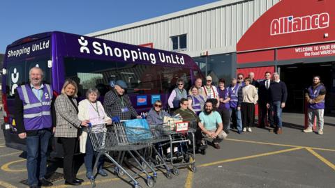 A purple minibus with white Shopping UnLtd branding on it. It is parked outside the door of the supermarket Alliance. Standing and in wheelchairs alongside the minibus with empty trolleys are a variety of people using the service wearing sunflower lanyards. Volunteers are wearing purple tabbards.