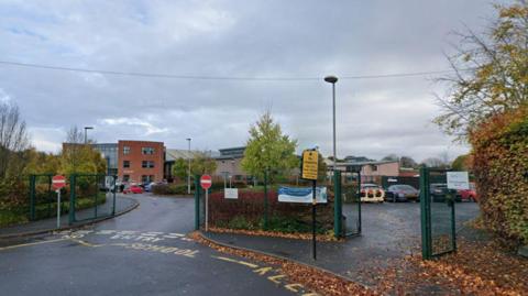 the outside of a secondary school, pictured from gates by its entrance. Green metal fencing bars an entrance and exit and a carpark can be seen in front of buildings. Hedges and trees line the perimeter. The school buildings can be seen in the distance, with three storeys. The sky is grey and cloudy.
