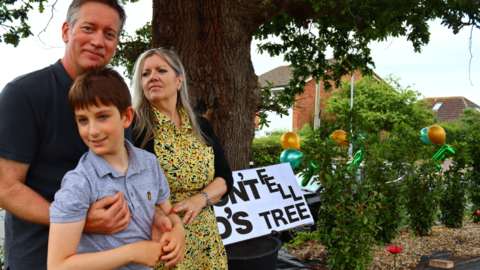 Man, woman and son standing in front of a large tree, with a sign next to it reading "DON'T FELL JO'S TREE,", The scene includes greenery, balloons, and a building in the background, suggesting a public or residential setting. 