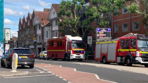 Two fire engines parked inside a cordon, with blue and white police tape securing a large section of the main road.