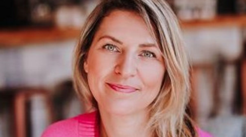 A woman with short blonde hair, she is slightly smiling and wears a pink cardigan.