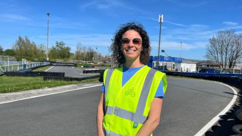 A lady in a high viz yellow jacket stands on a tarmac road. She's wearing sunglasses with a dark blue sky in the background. 