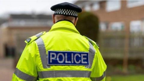 A police officer is standing with a high-vis jacket. He is looking away from the camera.