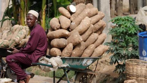 A yam seller dressed in his traditional attire with a cap sits on his wheel barrow containing piles of yam