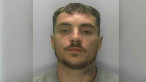 A police custody image of Tyler Doyle. He has short brown hair, a moustache and a beard. He has a tattoo on the left side of his neck.