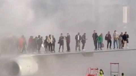 People stand on the wing of a plane on the tarmac. Smoke can be seen encircling around them, whilst airport staff on the ground begin to bring ladders to help them dismount.