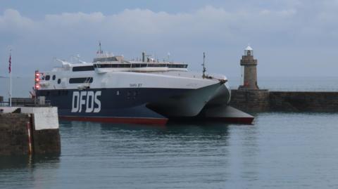 A wide shot of the DFDS coming into Guernsey Harbour. The sky is overcast. The boat is two tone, navy and white.