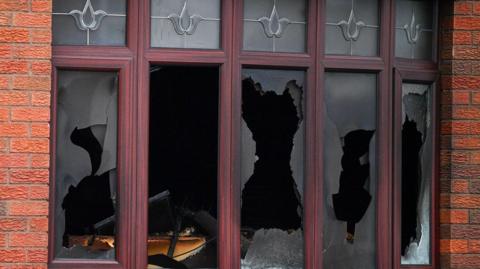 A house's front windows, most of which are smashed. The house's orange brickwork is visible around the windows.