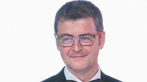 Brendan Mooney wearing a pair of glasses, a black jacket and white shirt. He is stood in front of a white background. 