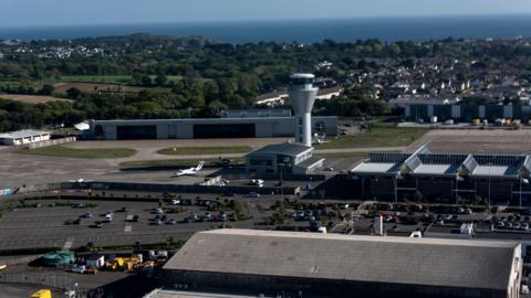 An aerial view of Jersey Airport with the full car park and building in vision, buildings fields and the sea further afield.