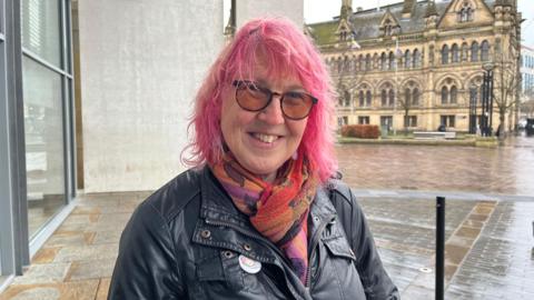 Sue Wilde, standing in front of City Hall in Bradford. She has pink hair and is wearing a black leather jacket. 