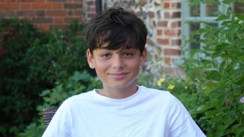 Kaan a boy with brunette hair who is sitting outside in front of a bush. He is wearing a white T-shirt and is looking directly at the camera and smiling.