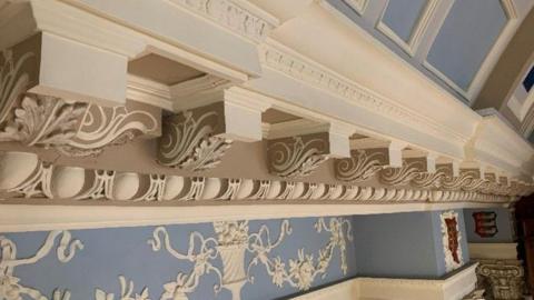 Part of an ornate ceiling. It is mainly blue and cream with plasterwork flowers in vases inlaid