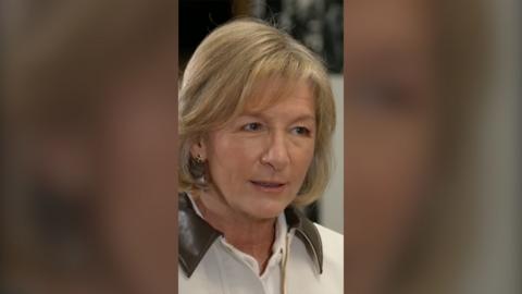 A woman with shoulder length blonde hair, wearing a white jacket with a brown collar