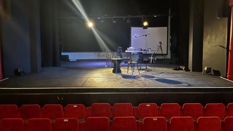 Rows of red seats in a theatre sit in front of a black stage which has a table and chairs along with theatre equipment