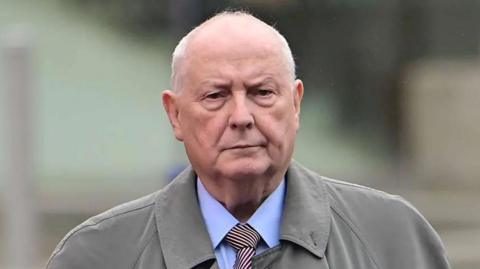 Mr Cushnahan is wearing a grey coat over a blue shirt and red and white stripped tie. He has short grey hair on his hair. The background is blurred. 