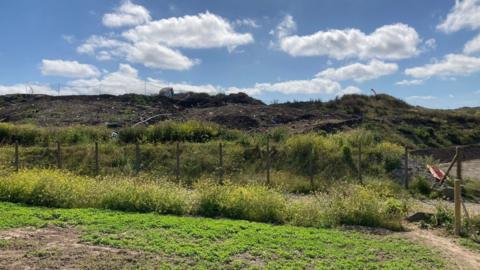 A landfill site showing a mound of dirt littered with rubbish including a boat. It is a partly cloudy day.