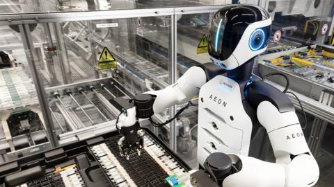 The Aeon robot at work in the factory. It has a white torso with black shoulders. Its black plastic face has a vertical line of blue light.