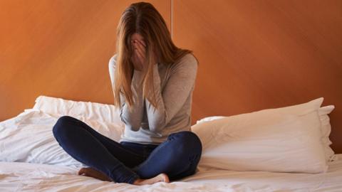A woman with long hair with her head in her hands on a bed with pillows behind her