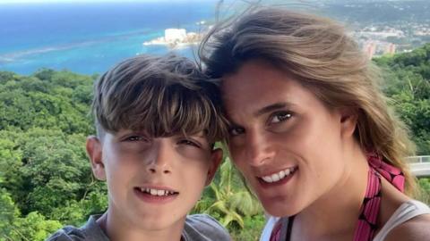 Ellen Roome with her 14-year-old son Jools Sweeney in a holiday snap. Behind them is vibrant green forest with a very blue sea in the background. Ellen has long brown hair and brown eyes and Jools has short brown hair. They are smiling at the camera.