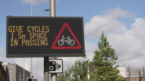 A digital road sign urging motorists to allow a 1.5m gap when passing cyclists. There is a red warning triangle with an image of a bike inside it. There are also trees and buildings in the background.