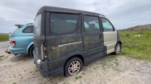 A weathered and neglected black van with moss and rust on the body panels, missing tail lights and a flat rear tyre parked on a dirt car park near grassy dunes. The sky above is a leaden grey colour with a blue SUV parked behind the van. 