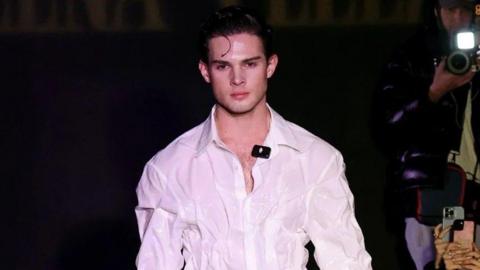 Braden Peters stares straight ahead as he walks a runway in a white button-up shirt.