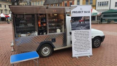 A truck with the side open showing lots of breakfast foods in the middle of a town square. 