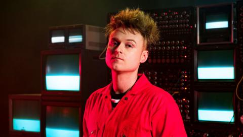 Sam Battle (Look Mum No Computer) wearing a red shirt standing in front of electronic equipment