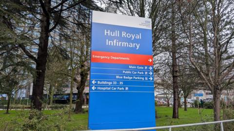 An image of Hull Royal Infirmary's main sign, with the hospital itself in the background behind a landscaped garden area.