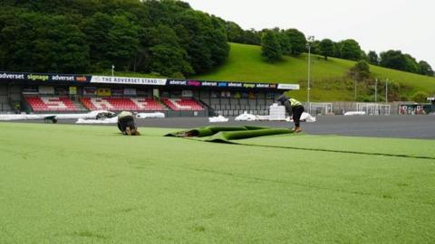 The image shows a football stadium undergoing resurfacing work. In the foreground there is a section of  green artificial turf, with two workers handling a rolled section. A long strip of turf is midway through being unrolled. Behind the workers is the main playing surface area. White bundles are scattered around the edges. To the right of the image a machinery arm is visible.