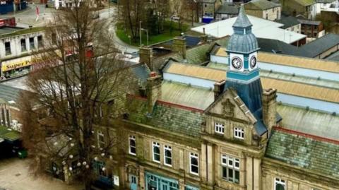 Darwen Town Hall's blue clocktower rising above a building with a crest on its gable. The photo is taken from above. A tree is next to the building with some lanterns in it and Super Poundland can be seen in the distance.