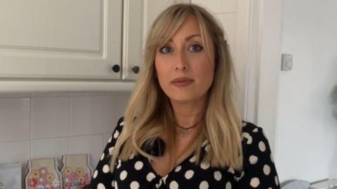 A woman with blond hair, wearing a black and white spotted top looks towards the camera. There are white kitchen cabinets behind her.