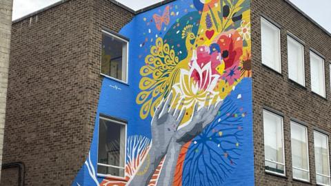 A closer view of mural on the side of a brick building. It depicts a person's hands that are open in offering with various flowers coming from them against a blue background. The building has several windows across multiple storeys. 
