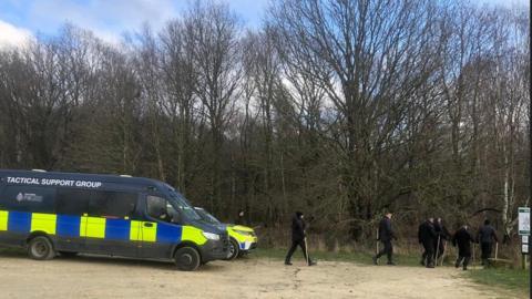 Police officers head into Wombwell Woods to search the woods and lake during, Tactical Support Unit van and police vehicle on the left. Seven police officers dressed in black carrying sticks head into the woods across the centre and right of the picture.