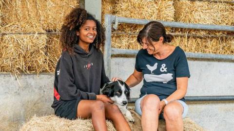 Amani (left) and Julie sit either side of Bella the dog on a hay bale. Amani is wearing a grey, red and black hoodie, black shorts and is smiling at the camera. Julie is wearing a T-shirt with a chicken and pig on it and denim shorts. Both are stroking Bella, who is black and white.