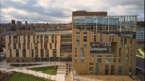 The University of Huddersfield's new Daphne Steel Building, on the National Health Innovation Campus (NHIC). A modern brown and gold building with windows and a triangle of grass and paths in the foreground. Huddersfield city centre can be seen in the distance.