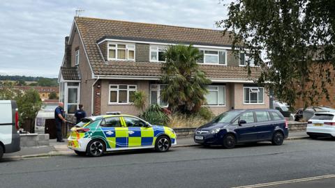 Two police officers in uniform standing outside a home in South Liberty Lane during the daytime, following the incident. It is a pebbledash building with slate roofs and a large palm tree outside. There are cars parked beside the pavement, including a police car with hi-vis blue and yellow decals. 