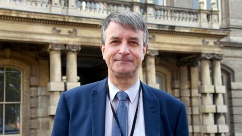 A head and shoulders shot of Councillor Philip Jackson. He has short grey hair, a light-coloured shirt, a navy blazer jacket and a light blue patterned tie. He is also wearing a black lanyard. He is stood in front of a building with stone columns.