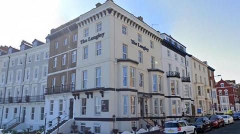 The Langley Hotel catches the eye on the corner of the street with its white walls, black decor, and its name in black on the front and side.  There are cars parked outside and a blue sky above.